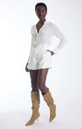 A model wearing a white long-sleeve collared shirt and matching white shorts with a gold belt, paired with tan knee-high heeled boots, against a plain white studio background.
