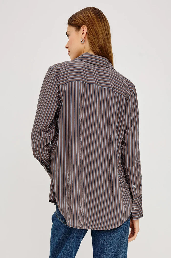A woman shown from the back wearing a long-sleeve button-up shirt with vertical blue, brown, and white stripes, paired with blue jeans, against a plain light gray studio background.