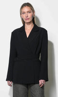 A woman modeling a black wrap-style blazer with a self-tie waist belt, paired with gray textured trousers, against a plain off-white background.