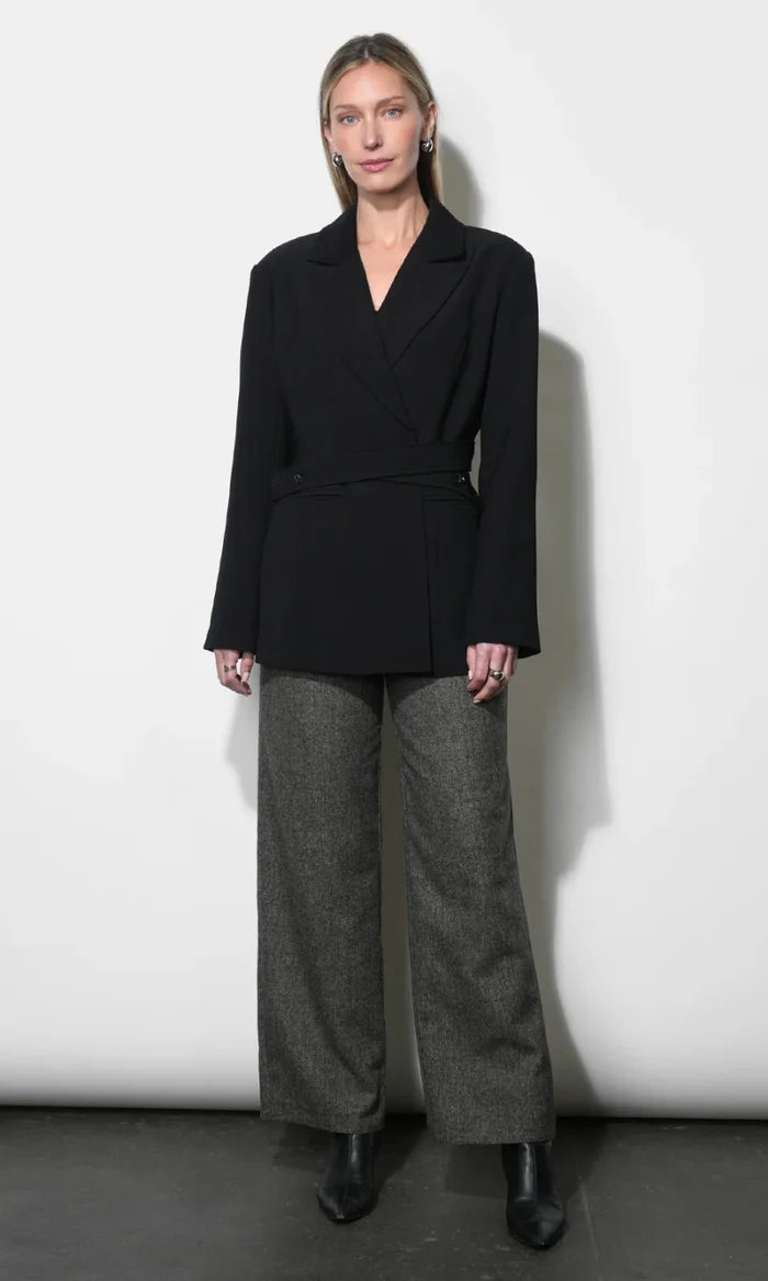A model stands against a white studio wall wearing a black belted wrap blazer with wide long sleeves, paired with wide-leg charcoal gray trousers and black ankle boots.