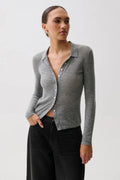 A woman in profile wearing a fitted gray ribbed knit polo cardigan with a buttoned collar over a black top and black pants, photographed against a light gray studio background.