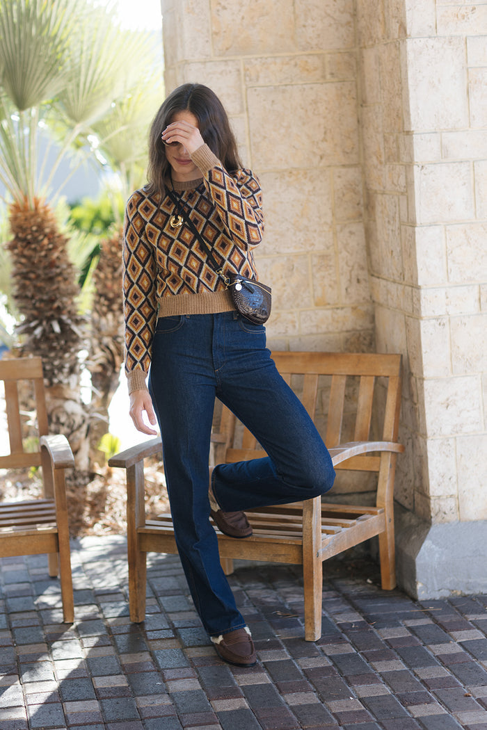 A woman posing outdoors in a brown and beige diamond-patterned knit sweater, dark blue jeans, and brown shoes, carrying a small black crossbody bag.