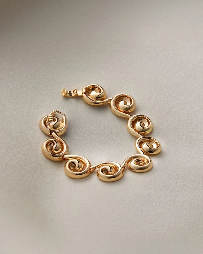A gold-plated link bracelet with a unique spiral or scroll design on each link, displayed on a neutral light gray background.