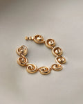 A gold-plated link bracelet with a unique spiral or scroll design on each link, displayed on a neutral light gray background.