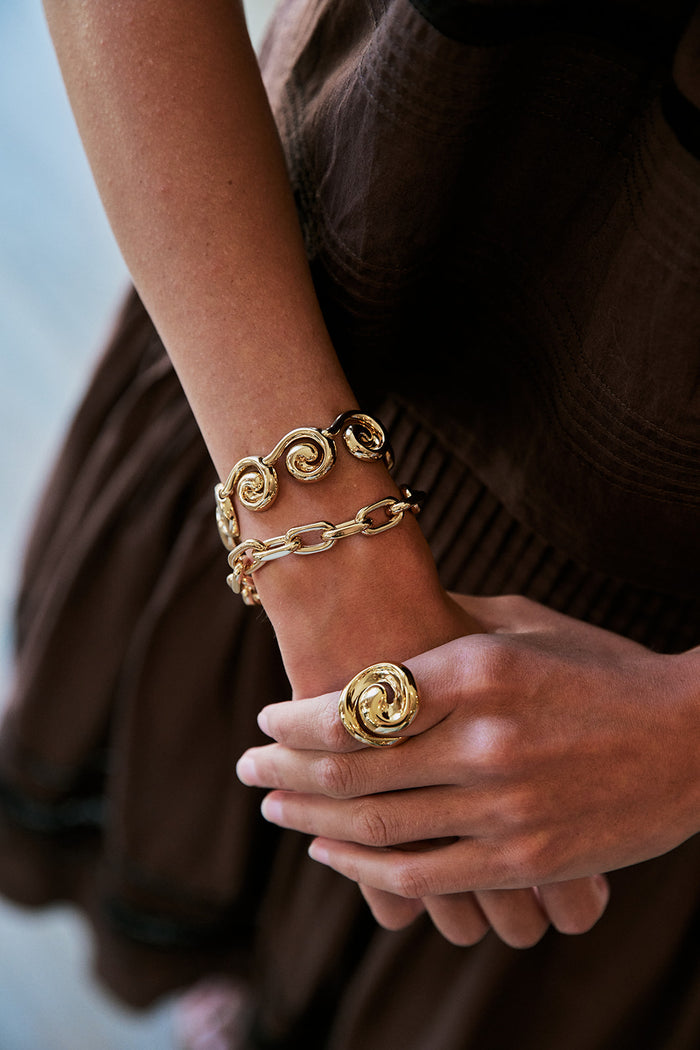 A close-up of a person's wrist and hand wearing multiple gold-tone jewelry pieces, including a spiral ring, a chain bracelet, and a bangle, against a dark brown dress.