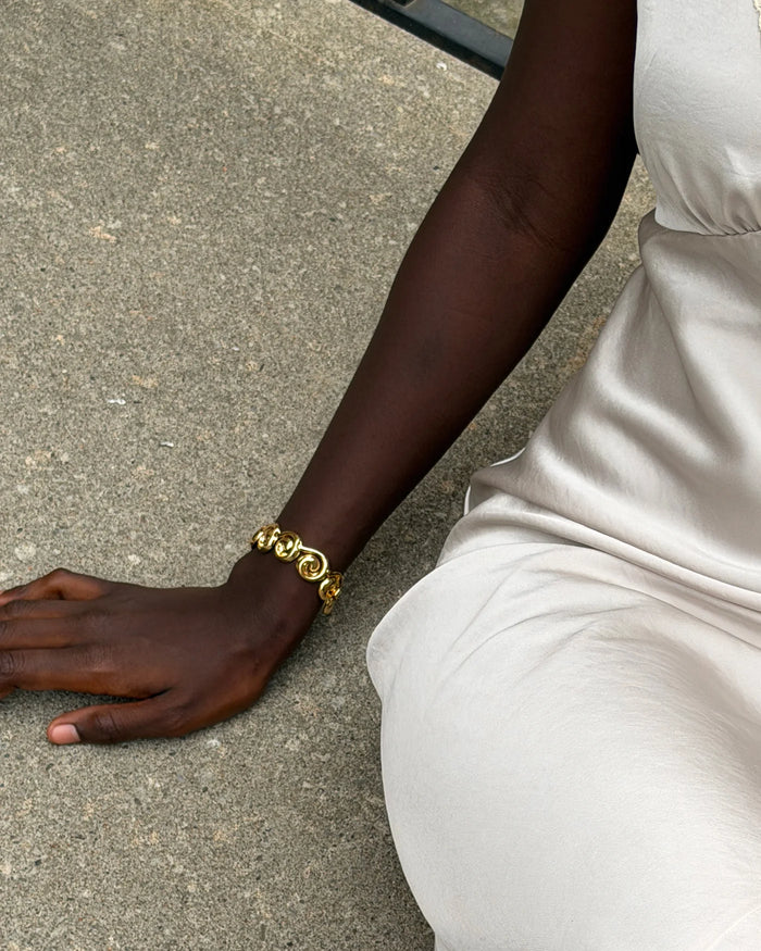 A close-up lifestyle shot of a person wearing a chunky gold chain link bracelet on their wrist, sitting on a concrete surface.