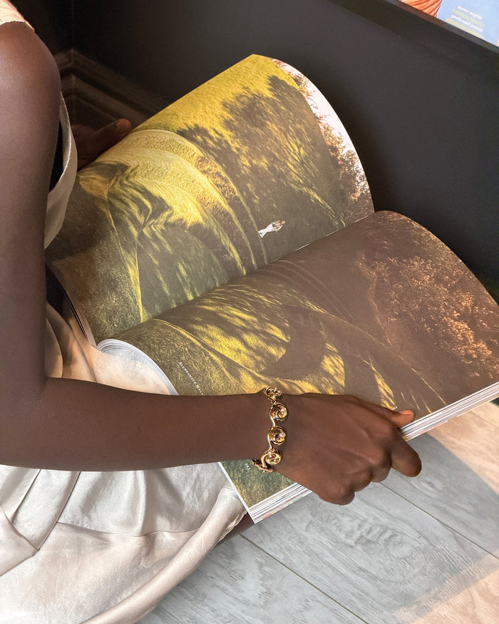 A person holding open a large printed book or magazine showing a landscape photograph with yellow and brown tones, wearing a gold chain bracelet on one arm.