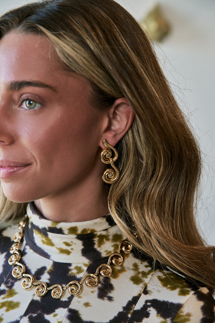 A close-up lifestyle shot of a woman wearing a matching gold spiral drop necklace and matching spiral drop earrings, paired with a high-neck tie-dye top.