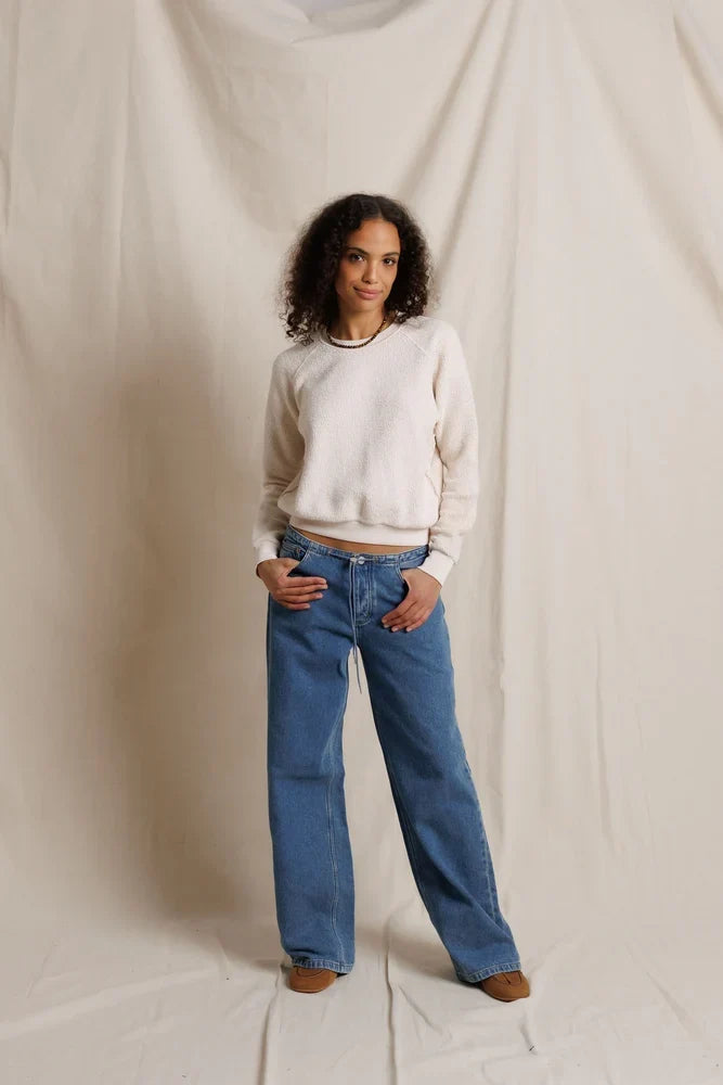 A woman stands against a draped cream fabric backdrop wearing a cream crewneck sweater, blue wide-leg jeans, and brown shoes.